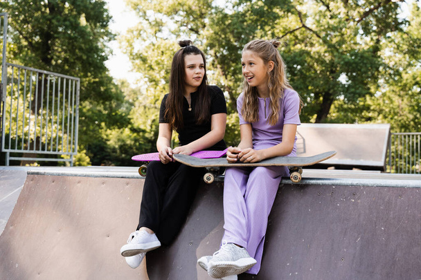 Sporty children girls with skateboard and penny boards are sitting and chatting with each other on sports ramp on playground. Friends communication and friendship. Sports extreme lifestyle - Foto, Bild
