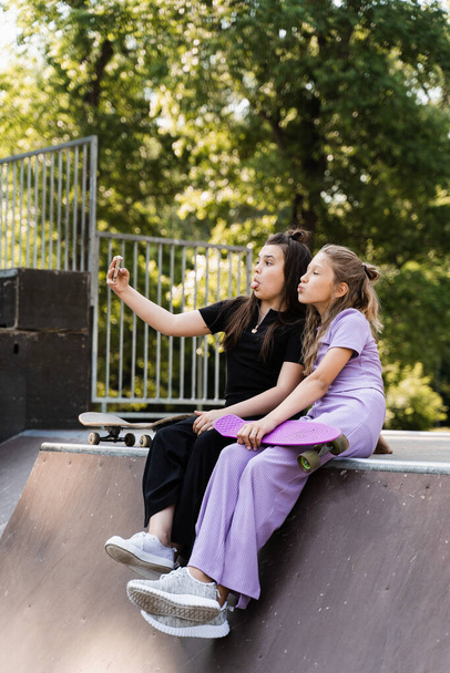 Phone addicted sports children with skateboard and penny boards use phones and making selfie instead of skating and play together. Children looking at smartphones on sports ramp. Phone addiction - Photo, image