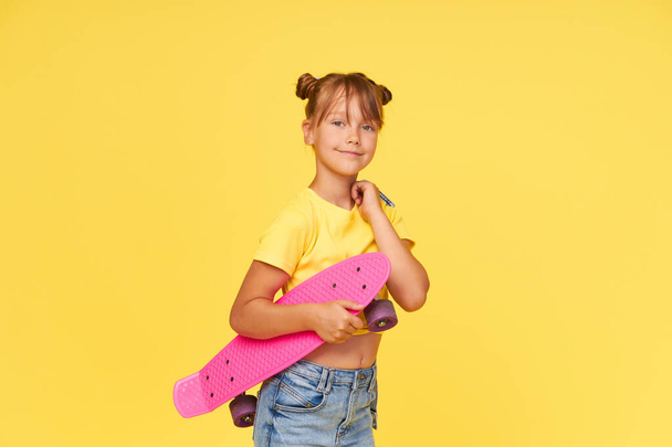 A cute little girl 6 years old in a bright tulle skirt and a white t-shirt and stylish glasses holds a bright colored skateboard on a yellow background - Zdjęcie, obraz
