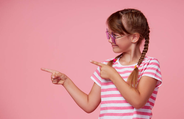 Portrait of surprised cute toddler girl child in star-shaped sunglasses over pink background. Points hands to the left side. Advertising childrens products - Foto, afbeelding