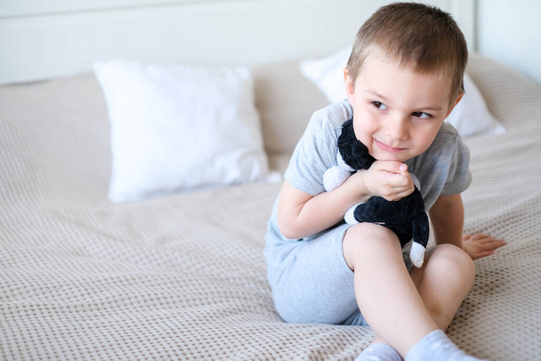 Close-up of a small happy caucasian boy in a gray t-shirt lies on the bed with a toy dog. - Фото, зображення