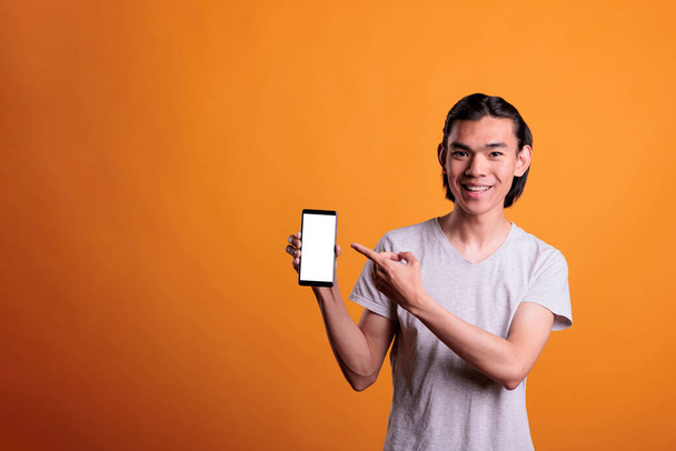 Young asian man pointing at smartphone blank screen with copy space, teenager holding mobile phone with place for text. Smiling person presenting telephone with empty display, mock up for promo - 写真・画像