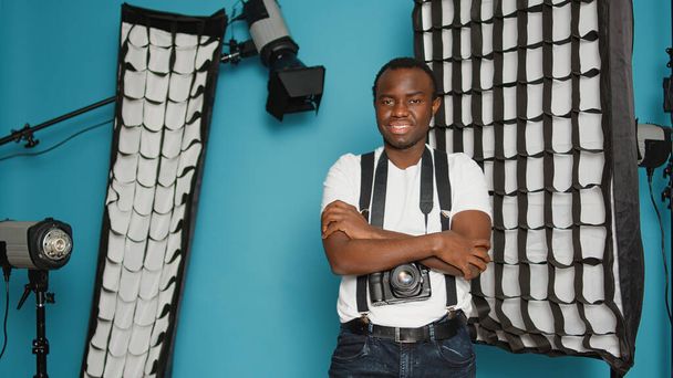 Male photographer posing with arms crossed in studio backstage, having professional photography equipment. Using behind the scenes lighting, reflector, tripod and softbox tools. - Photo, Image