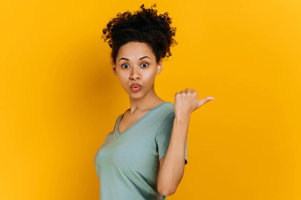 Удивлённая эмоция африканская кудрявая американская молодая женщина в футболке, указывает пальцем в сторону за спиной, на пустое пространство для копирования, стоит на изолированном оранжевом фоне, потрясенный взгляд на камеру - Фото, изображение