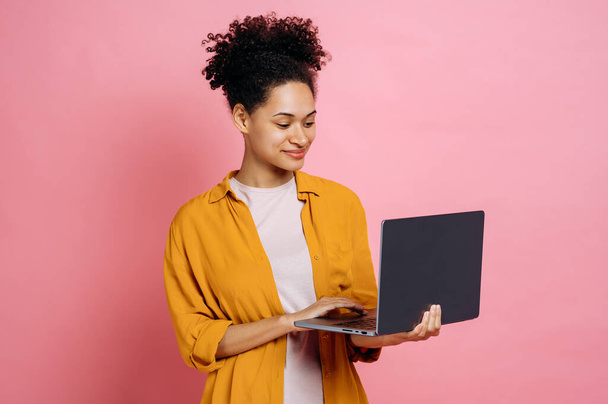 Θετική όμορφη Αφροαμερικανή μοντέρνα νεαρή σγουρομάλλα γυναίκα, κρατώντας ένα ανοικτό φορητό υπολογιστή στο χέρι, κοιτάζει την οθόνη, στέκεται απέναντι σε απομονωμένο ροζ φόντο, χαμογελώντας ευτυχισμένοι. Ασύρματη τεχνολογία - Φωτογραφία, εικόνα