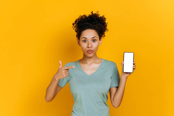 Shocked stunned african american curly young woman, surprised looks at the camera, holds cell phone with empty white screen and points finger at it, stand on isolated orange background - Foto, immagini