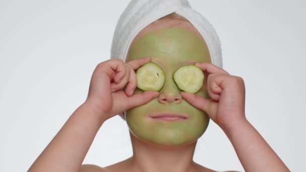Smiling child girl applying cleansing moisturizing green facial mask, holding slices of cucumber, covering eyes. Teenager kid face skin care treatment natural cosmetics. Female close-up macro portrait - Imágenes, Vídeo