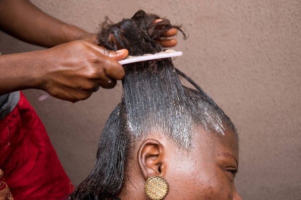 Cabeleireiro relaxando o cabelo em uma cabeça de mulher africana e também usando pente para esticar e aplicar o creme relaxante através do cabelo - Foto, Imagem