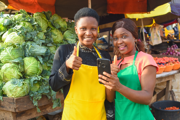Две счастливые африканские деловые женщины или продавщицы носят яркие фартуки, стоя в овощной ларьке на рынке со смартфоном. - Фото, изображение