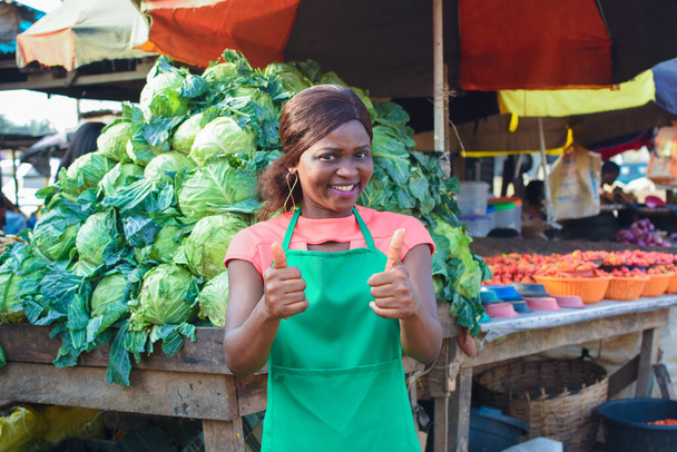 Happy African business woman or female trader wearing a green apron, doing thumbs up gestures while standing at her stall of vegetables in a market place - Fotoğraf, Görsel