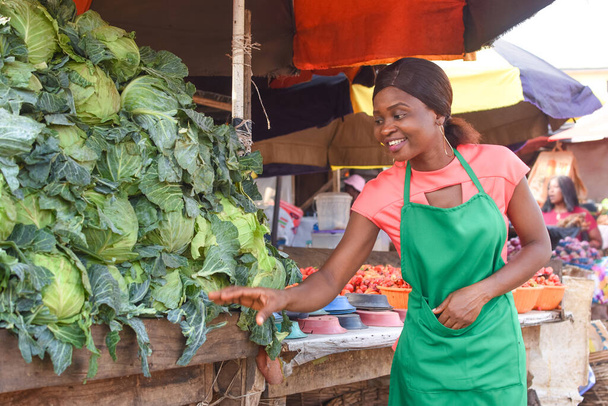 Heureuse femme d'affaires africaine ou commerçante portant un tablier vert et s'occupant de ses marchandises de légumes sur une place de marché - Photo, image