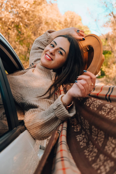 Brunette European looking cute girl sitting in a car enjoying sunny autumn weekend. Travel and road trip. Woman looking at the view from a car. Happy woman enjoying road trip. Fall Autumn forest in - 写真・画像