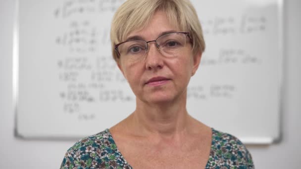 Aged concentrated blonde woman lecturer with poor eyesight in glasses and dress front portrait. Whiteboard with equations and algebra materials on the background. - Séquence, vidéo