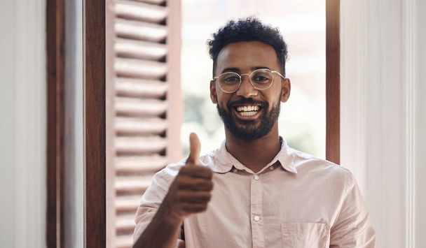 Thumbs up, endorsing and thank you with a young man smiling, feeling positive and giving his approval while standing inside. Portrait of a happy male wearing glasses and giving his trust or support. - Photo, Image