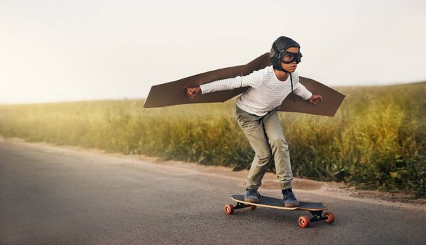 Hes got the wildest imagination. a young boy pretending to fly with a pair of cardboard wings while riding a skateboard outside - Фото, изображение
