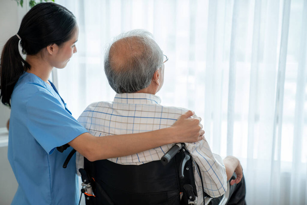 Rear view of a caregiver and her contented senior patient gazing out through the window. Elderly illness, nursing homes for the elderly, and pensioner life - Foto, Imagen