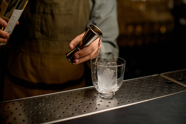 hand of male bartender carefully holds steel jigger and pours drink from it into a transparent glass with an ice cube on bar - Zdjęcie, obraz