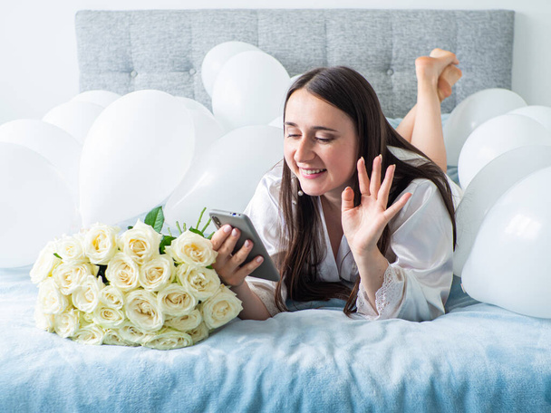 Женщина в белом лежит между белыми шариками на кровати. Портрет улыбающейся женщины с помощью телефона с большим количеством белых воздушных шаров и букета роз. С днем рождения, свадьба. Общение онлайн. - Фото, изображение