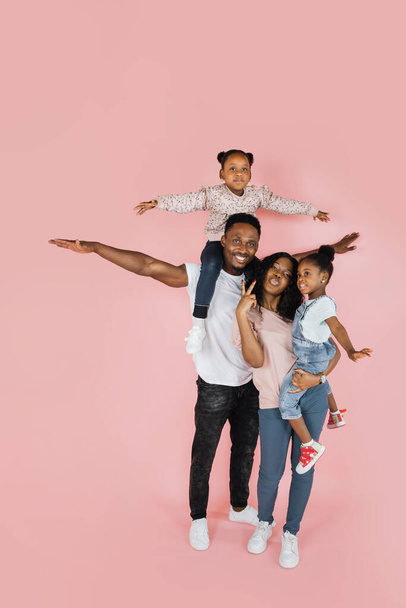 Family bonding concept. Joyful black man and woman holding daughters on hands, having fun over pink studio wall. Smiling girls pretending they are flying, spreading hands, imitating plane - 写真・画像