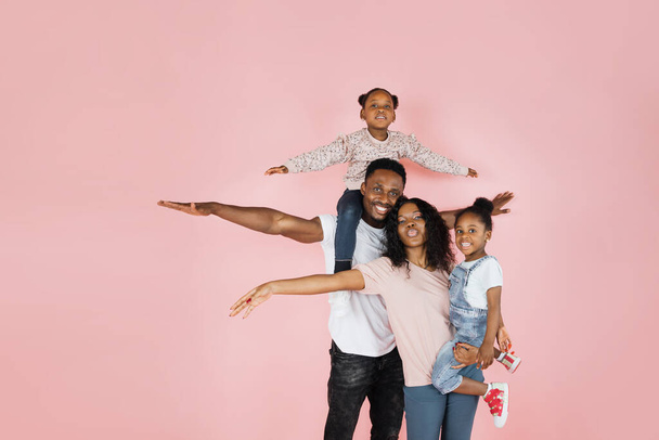 Family bonding concept. Joyful black man and woman holding daughters on hands, having fun over pink studio wall. Smiling girls pretending they are flying, spreading hands, imitating plane - Valokuva, kuva