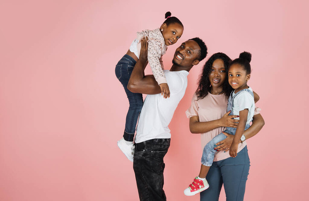 Time with family. Overjoyed african american family laughing and posing isolated over pink studio wall. Cheerful father and mom carrying their daughters on hands. - Fotó, kép