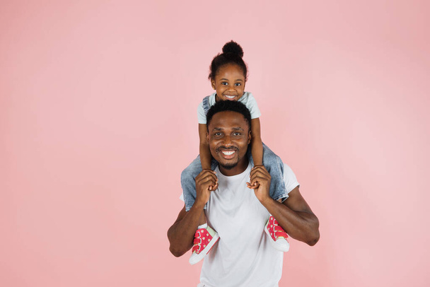 Happy family. Portrait of cheerful african american man riding excited daughter on his shoulders, looking and posing at camera, having fun, holding hands isolated over pink studio background. - Фото, зображення