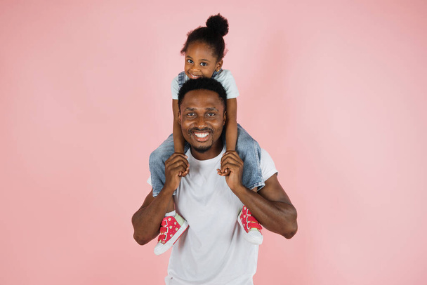 Happy family. Portrait of cheerful african american man riding excited daughter on his shoulders, looking and posing at camera, having fun, holding hands isolated over pink studio background. - Φωτογραφία, εικόνα