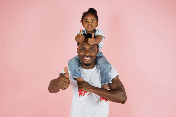 Portrait of cheerful african american man riding excited daughter on his shoulders showing thumbs up, looking and posing at camera, having fun, holding hands isolated over pink studio background. - Foto, Imagen