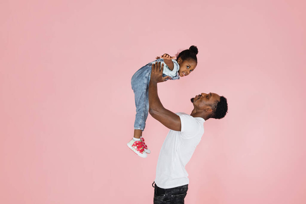 Happy fatherhood. Cheerful african dad holding his cute daughter on hands pretending she are flying, girl spreading hands, imitating plane, pink studio background. - Fotografie, Obrázek
