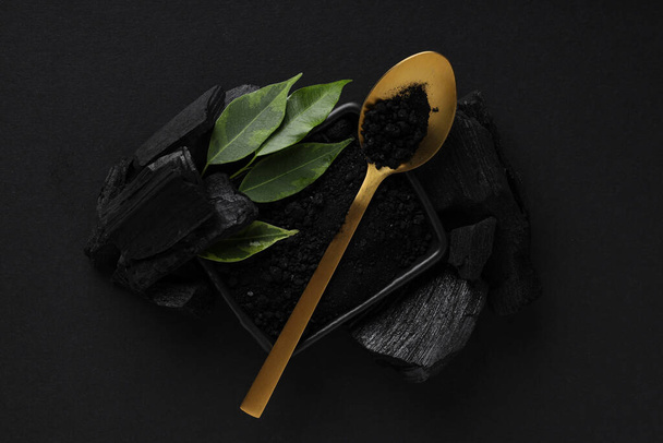 Charcoal, powdered charcoal, spoon and leaves on black background - Фото, изображение