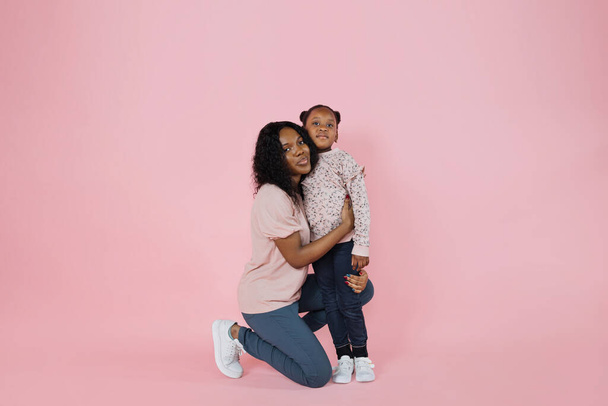 Indoor studio shot of pleasant young African woman mother and her cute adorable little daughter, wearing bright trendy casual clothes, posing together on pink background. - Foto, Imagen