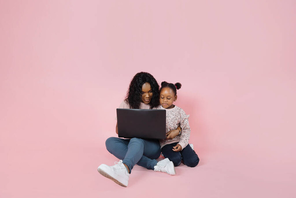 Happy african american mom and little cute daughter spending time together using laptop, sitting together on floor over pink background, copy space. - Photo, Image
