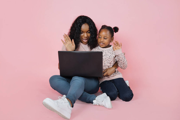 Horizontal shot of happy joyful African family, young mommy with cute little kid daughter, sitting on the floor on pink studio background and waving hands during online video call on laptop pc. - Valokuva, kuva