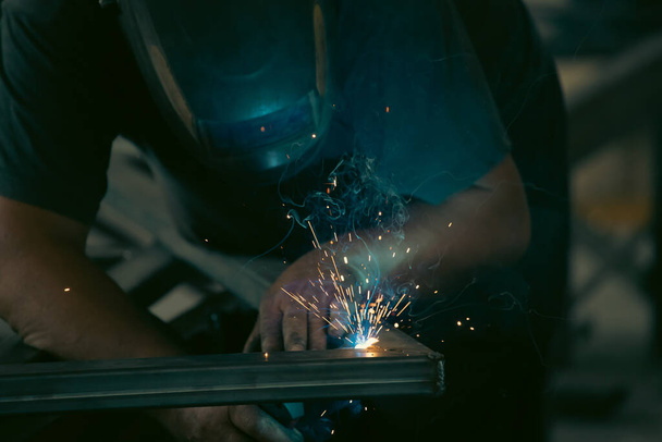 Handyman performing welding and grinding at his workplace in the workshop, while the sparks "fly" all around him. He is wearing a protective helmet and equipment. - Foto, Imagen