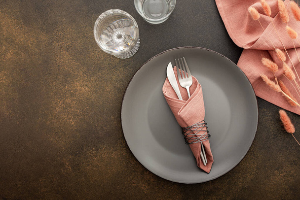 Asztalbeállítás, üres szürke tányér szalvétával és evőeszközökkel barna alapon, a felszolgált asztal tetejére néző, száraz virágokkal díszített - Fotó, kép