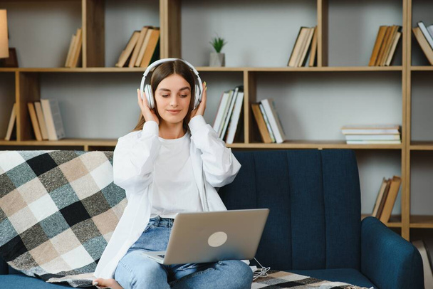 Beautiful young caucasian woman in casual clothing enjoying music and smiling while resting at home. Young woman with headphones using laptop at home. - Photo, image