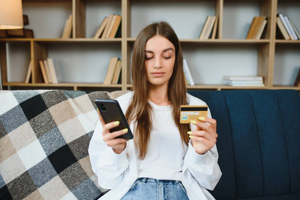 Happy teenager doing shopping online sitting on couch at home. Young woman insert credit card number on website with her smart phone - Fotó, kép
