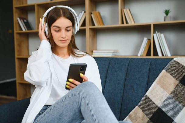 Beautiful young caucasian woman in casual clothing enjoying music and smiling while resting at home. Young woman with headphones using laptop at home. - Photo, image