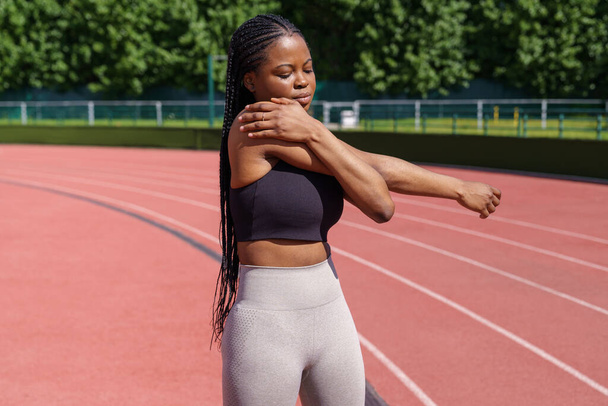 African American sportswoman with long braids enjoys training in sunny morning. Young woman stretches hands and shoulders standing on red track of stadium against lush green trees in city park - Photo, Image
