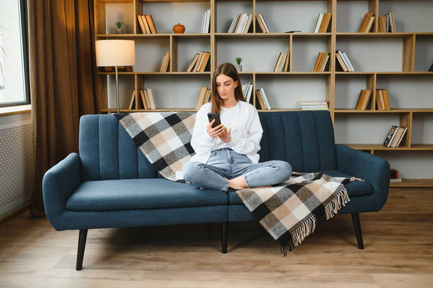 Young happy caucasian woman she 20s in casual clothes hold in hand use mobile cell phone sit on blue sofa indoor rest at home in own room apartment. People lifestyle concept. - Φωτογραφία, εικόνα