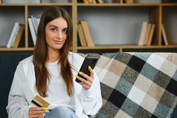 Happy teenager doing shopping online sitting on couch at home. Young woman insert credit card number on website with her smart phone - Foto, Imagen