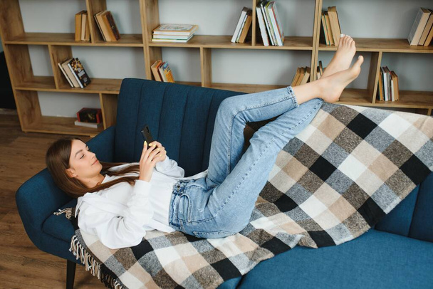 Young beautiful woman wearing white t-shirt on textile sofa at home. Attractive slim female in domestic situation, resting on couch in her lofty apartment. Background, copy space, close up - Φωτογραφία, εικόνα
