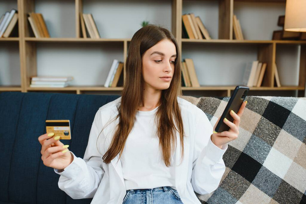 Happy teenager doing shopping online sitting on couch at home. Young woman insert credit card number on website with her smart phone - Фото, зображення