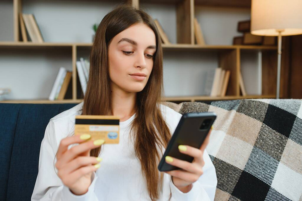 Happy young woman holding phone and credit card sitting on the couch. Online shopping - Fotó, kép