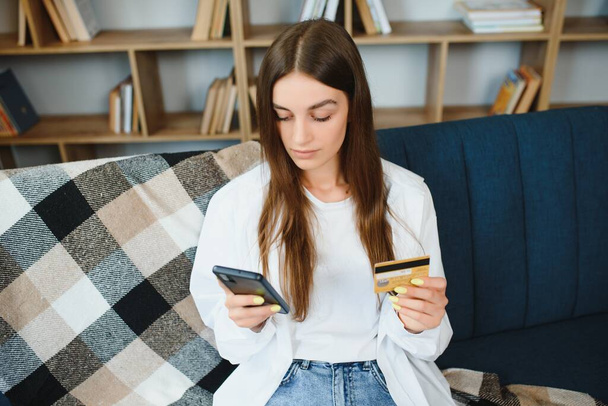 Adolescente feliz haciendo compras en línea sentado en el sofá en casa. Mujer joven inserte el número de tarjeta de crédito en el sitio web con su teléfono inteligente
 - Foto, Imagen