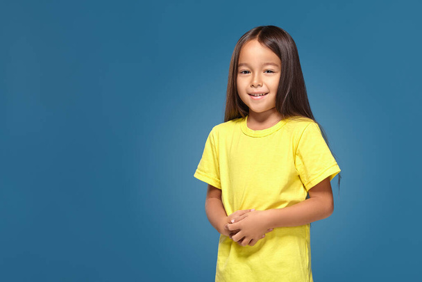 Το κοριτσάκι με το κίτρινο μπλουζάκι χαμογελάει σε μπλε φόντο. - Φωτογραφία, εικόνα