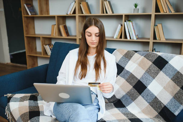 Smiling woman holding credit card and using laptop on sofa in living room - Foto, Imagen