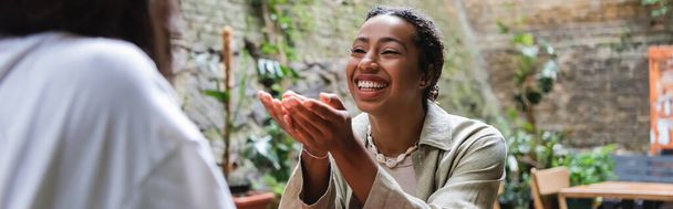 Позитивная африканская американка разговаривает с размытым другом в кафе на открытом воздухе, баннер  - Фото, изображение