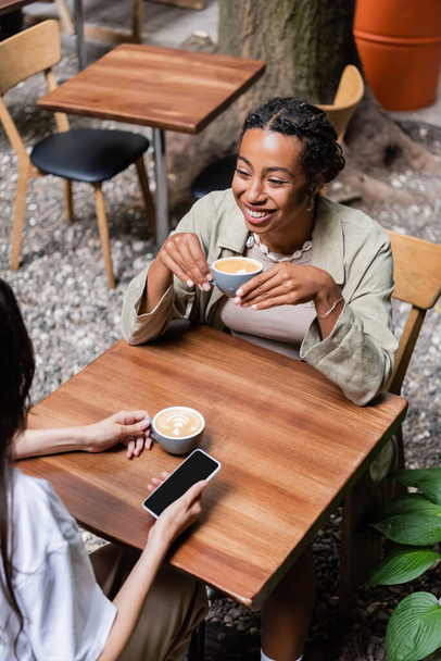 High angle view of cheerful african american woman holding cup of coffee near friend with cellphone on terrace of cafe  - Photo, Image