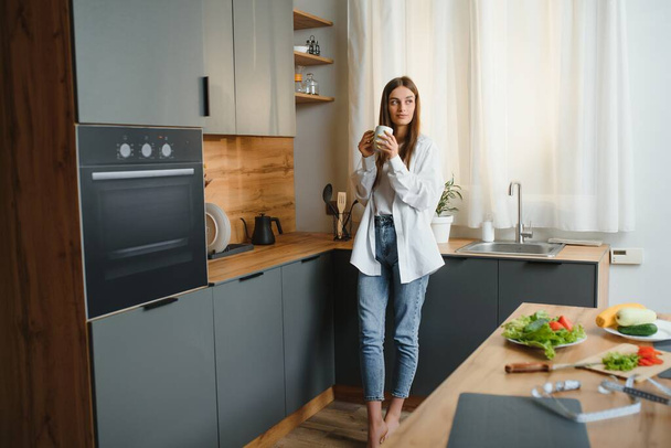 Happy young woman holding a cup of tea while standing in the kitchen at home in the morning. Healthy lifestyle concept - Foto, Imagen
